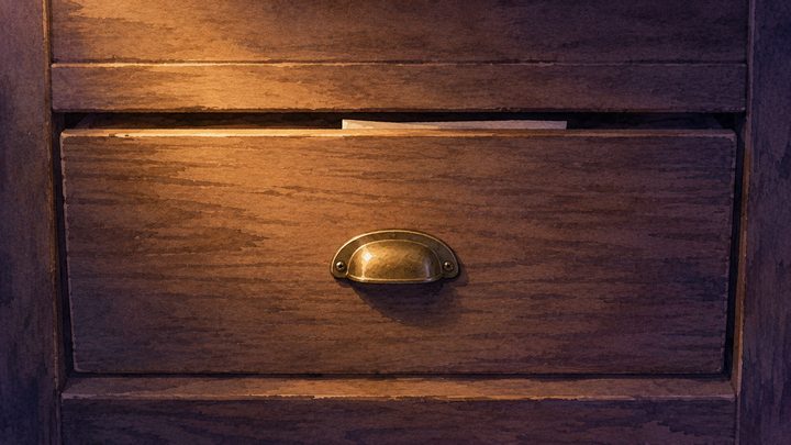 Overhead mid-shot at dusk of a closed wooden cabinet drawer with a thin tab of paper just visible at the seam
