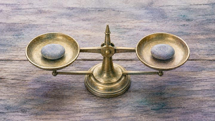 Close-up overhead of a small brass balance scale level on a wood surface, each pan holding an equal smooth stone, overcast light